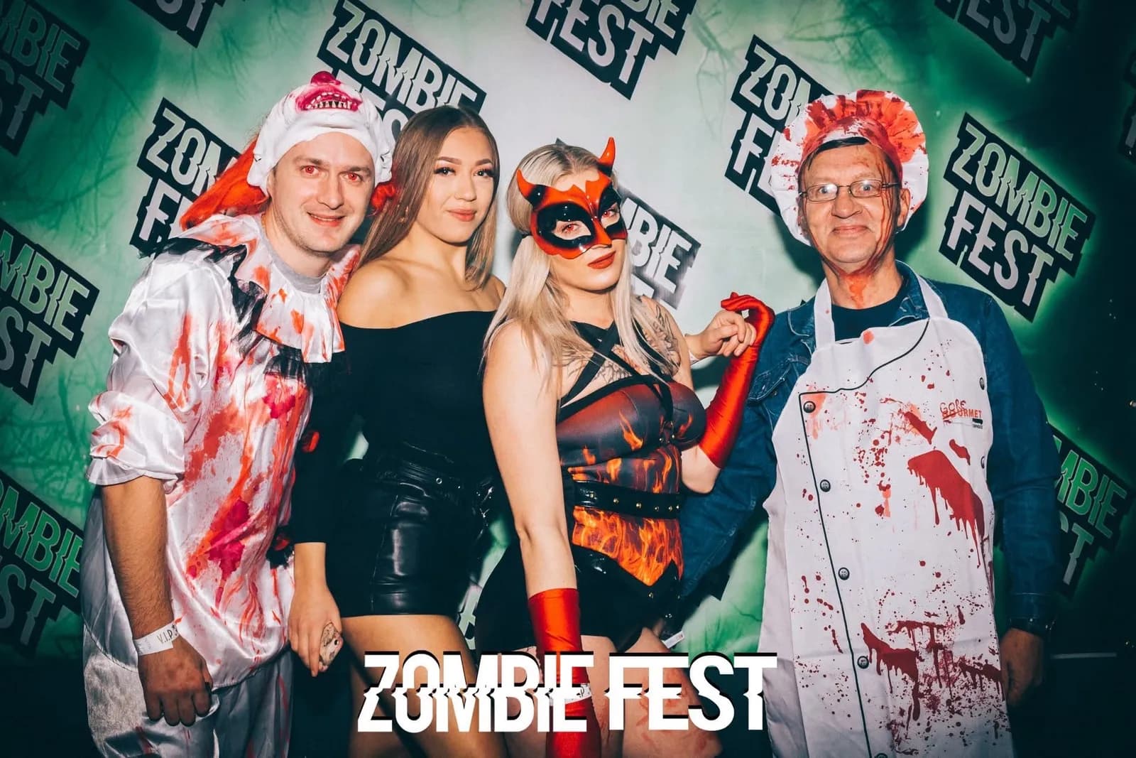 Four people dressed in zombie-themed costumes pose for a photo in front of a green backdrop with "Zombie Fest" written on it.
